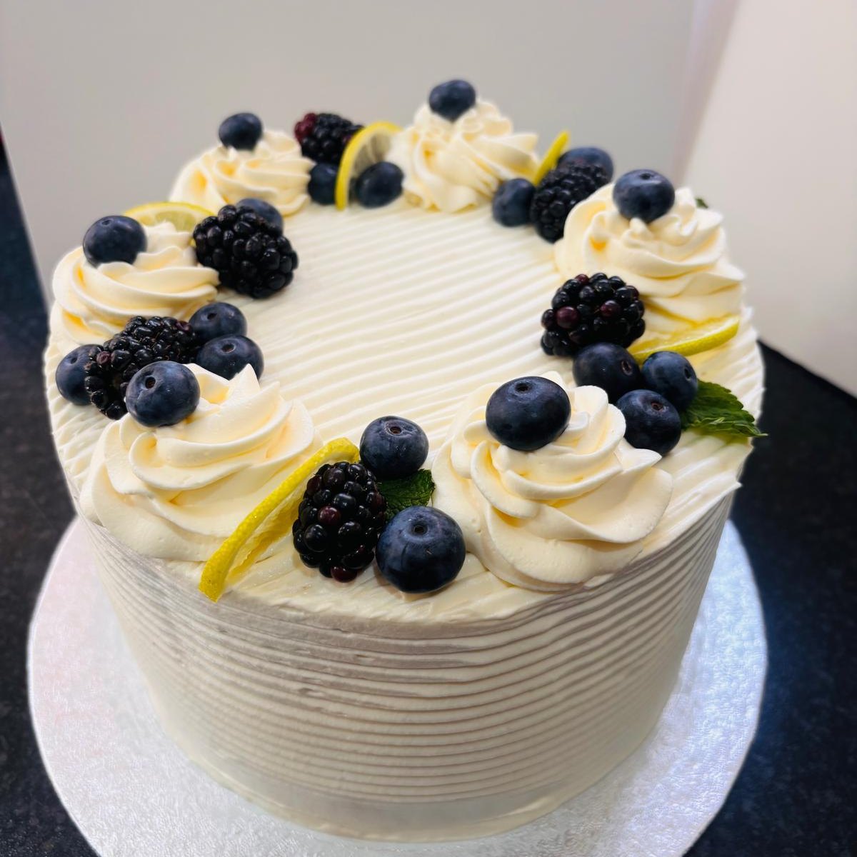A cream frosted cake ringed with piped rosettes, fresh blueberries, blackberries and lemon zest, baked in Medway